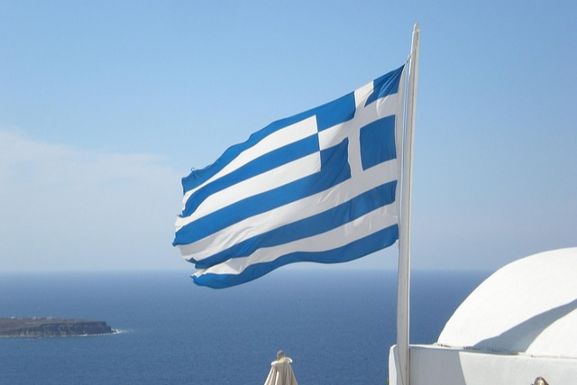 Griechische Flagge weht im Wind über dem Meer, im Hintergrund Küste und blauer Himmel – Symbol für Griechenlands Kultur, Meer und Sonne.