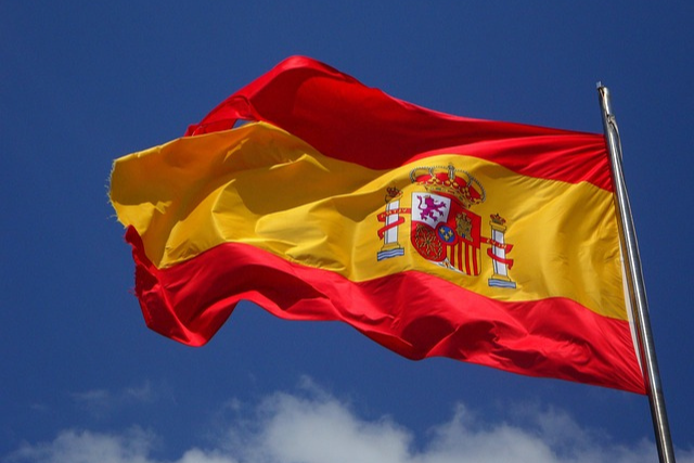 Spanische Flagge mit rotem und gelbem Streifen sowie Wappen, weht im Wind vor blauem Himmel – Symbol für Stolz und Kultur Spaniens.