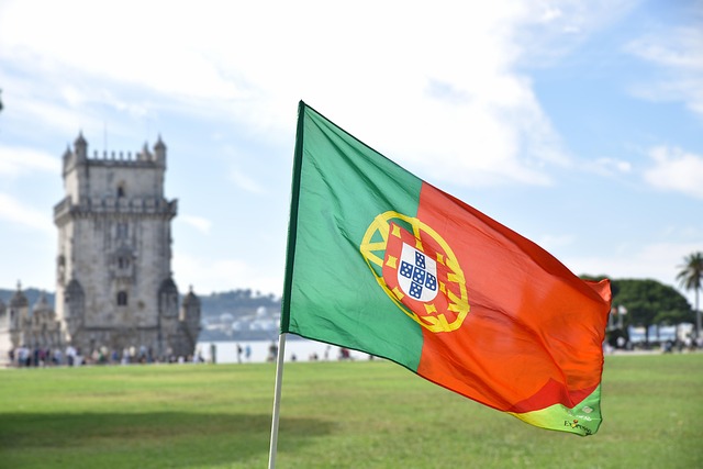 Portugiesische Flagge weht im Wind vor dem Belém‑Turm in Lissabon, historische Festung am Ufer des Tejo unter blauem Himmel – Symbol für Portugals Geschichte und Identität.